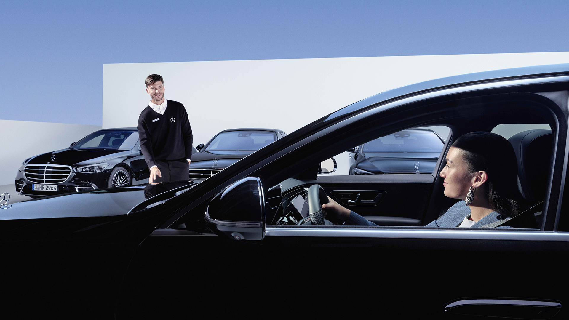Henkilö istuu mustassa Mercedeksessä. Taustalla seisoo asiakaspalvelija takanaan useita Mercedeksiä.