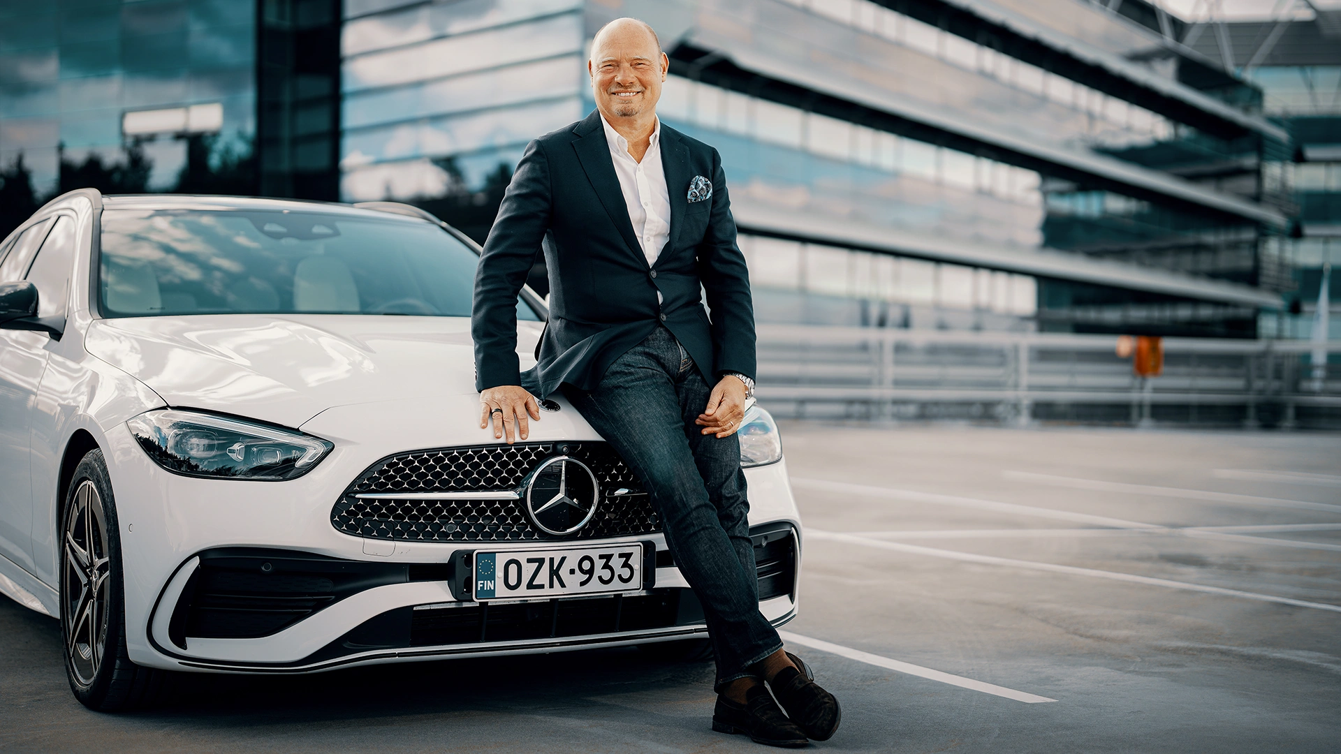 Mies istuu Mercedes-Benz henkilöauton keulalla toimistorakennuksen edessä.